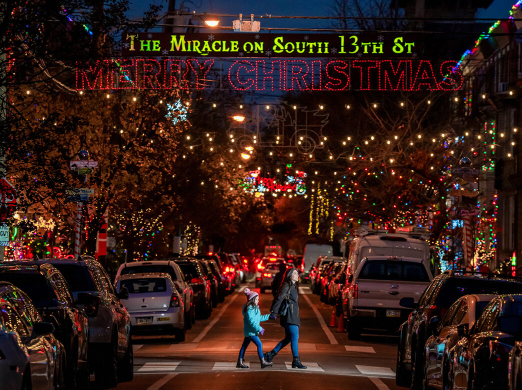 A mother and daughter hold hands while crossing South 13th Street during the Miracle on South 13th Street. Christmas lights decorate homes on the street.