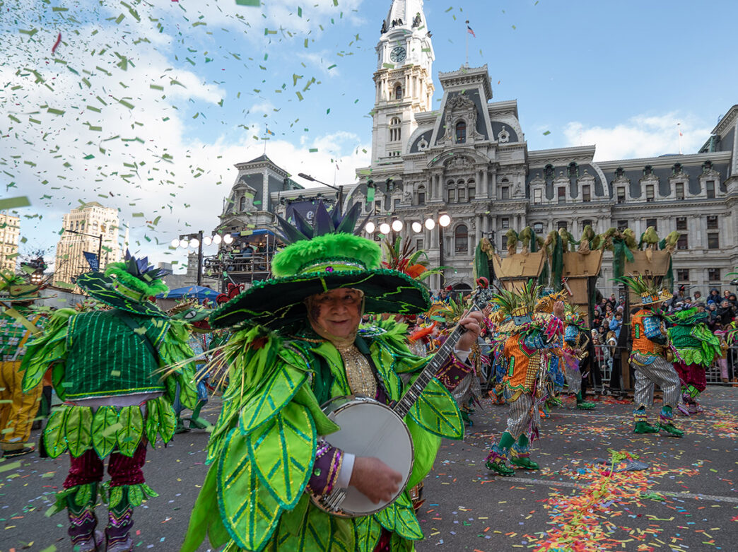 A performer dressed in vibrant green and gold feathers plays a banjo in front of Philadelphia City Hall during the Mummers Parade.