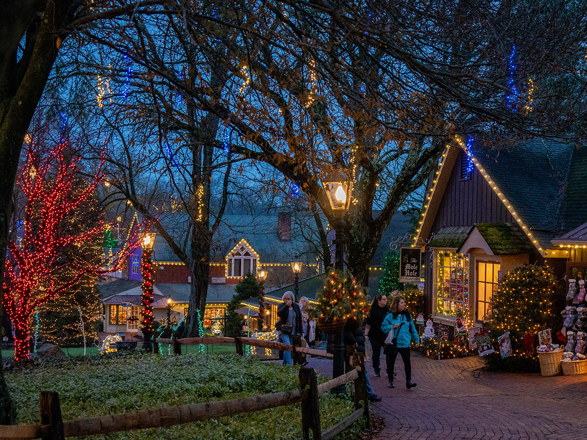 Festive holiday lights illuminate the shops and trees at Peddler's Village during a peaceful evening. Visitors stroll along the brick paths, surrounded by vibrant red, yellow, blue and green string lights and charming storefront decorations.