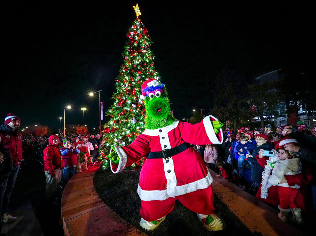 The green Phillie Phanatic mascot wears a red Santa suit and poses for a photo in front of a decorated Christmas tree. A crowd of people wearing Phillies hats and jackets circle around the tree.