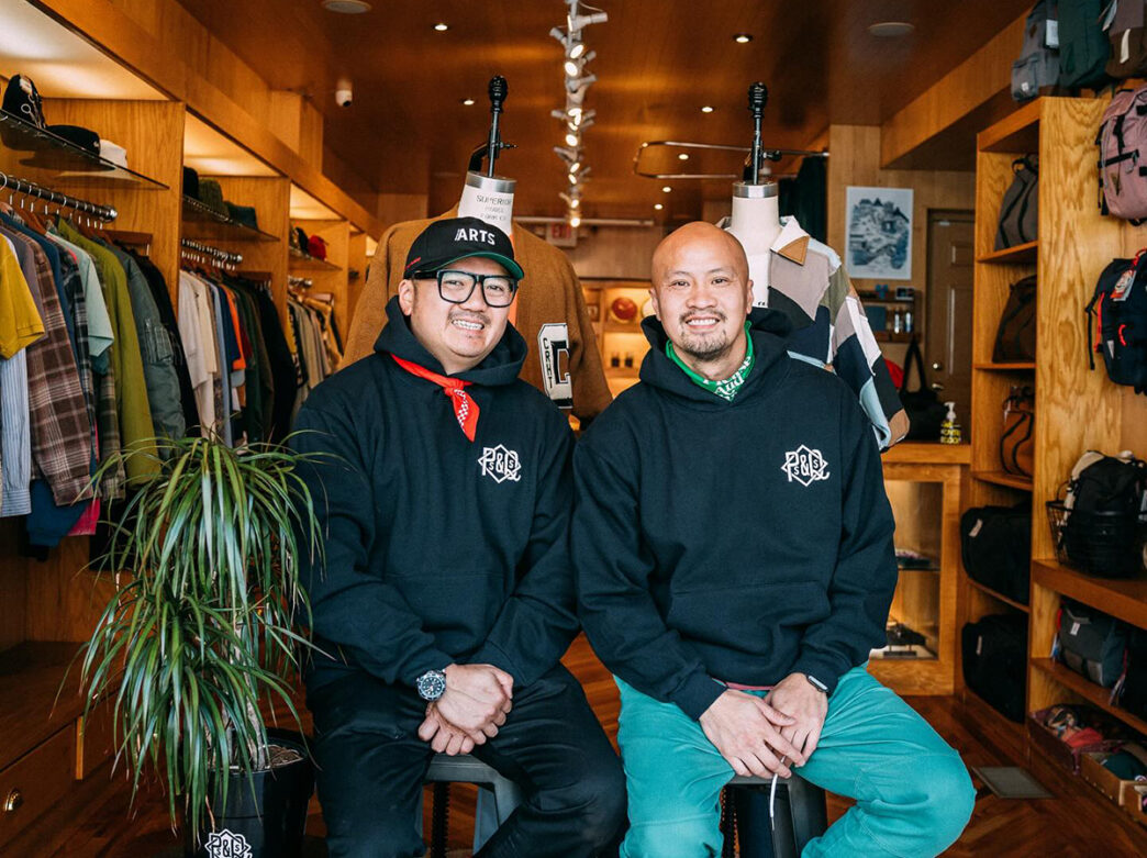 Rick and Ky Cao, owners of P's & Q's, sit on stools and pose for a photo together in their shop. Clothes hang on racks and bags are displayed on shelves behind them.