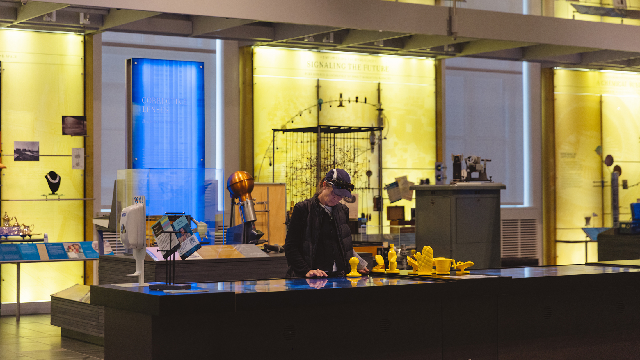 A person looks down at a digital screen at an interactive exhibit at the Science History Museum. Yellow and blue displays are in the background.