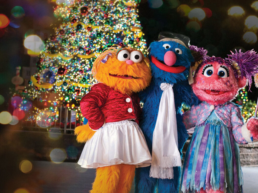Grover, Abby Cadabby, and Zoë from Sesame Street don festive holiday outfits and pose in front of a brightly lit Christmas tree at Sesame Place.