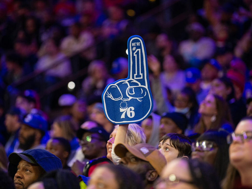 A lively crowd at a Philadelphia 76ers game in the Wells Fargo Center. One young fan holds up a blue foam finger that reads 'We're #1' alongside the team's logo.