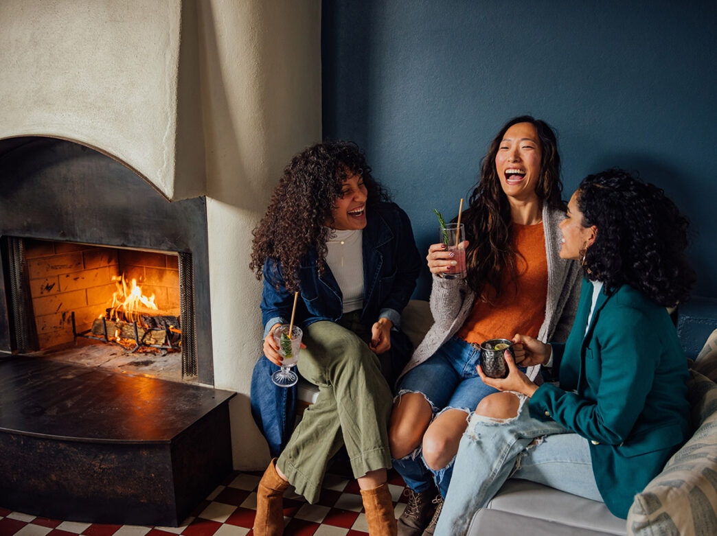 Three women laugh and enjoy drinks while sitting near a cozy fireplace in the warm and inviting ambiance of the restaurant Suraya.