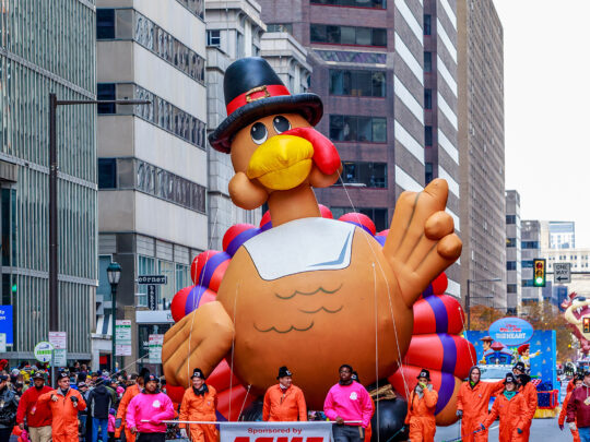 A large turkey float is marched down the street during the Thanksgiving Parade in Philadelphia. Crowds of people watch the parade from the sidewalks.