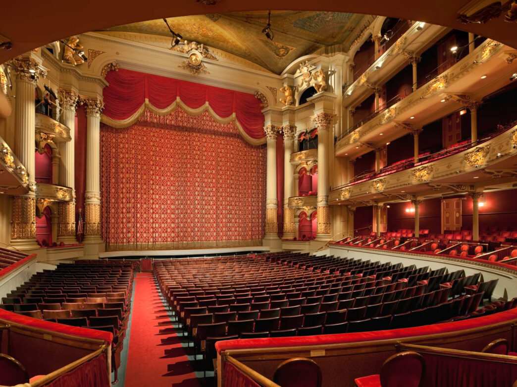 Una vista del interior del teatro de la Academia de Música. Un telón rojo cubre el escenario. El teatro está decorado con detalles en dorado y marfil, además de butacas de tela roja.