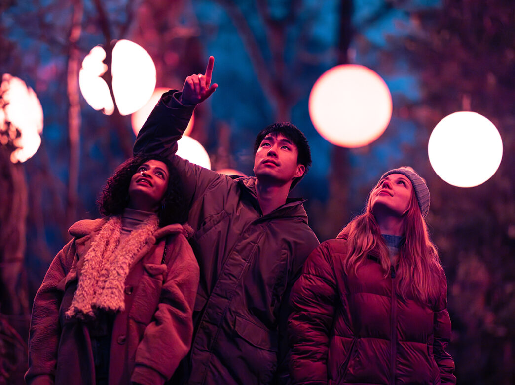 Three people wearing winter jackets look up towards the sky while one of them points upward. Large ball lights hang from trees around them.