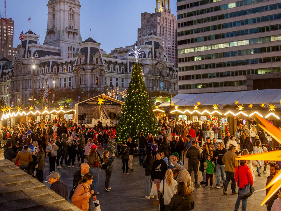 Vendor huts with decorative lighting and a large Christmas tree are set up in Love Park for Philadelphia's Christmas Village. People walk around shopping and taking photos. City Hall is in the background.