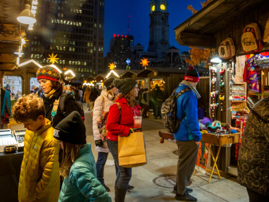 People wearing jackets and hats shop at night at the Christmas Village in LOVE Park. The vendor huts have lights to illuminate their goods.