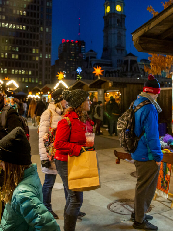 People wearing jackets and hats shop at night at the Christmas Village in LOVE Park. The vendor huts have lights to illuminate their goods.