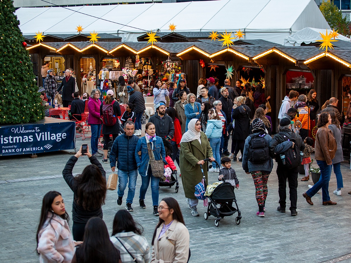 A crowd of people wearing jackets, hats and scarfs walk around and shop at the Christmas Village in Philadelphia. A Christmas tree and vendor huts can be seen in the background.