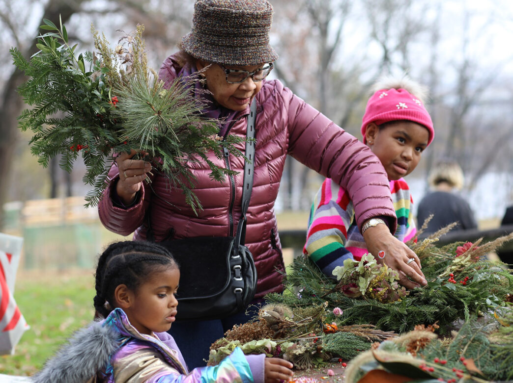 A person and two children look through twigs and greenery displayed on a table at the wreath-making station during Handmade Holidays at Bartram's Garden.