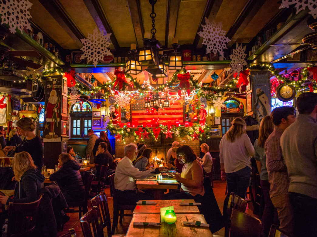 People sit at tables and stand at the bar in McGillin's Olde Ale House. The interior is decorated with garland, paper snowflakes and string lights.