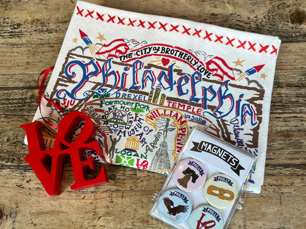 A red Love Statue ornament, a dish towel decorated with Philadelphia neighborhoods and a magnet set are displayed on a wooden table top.