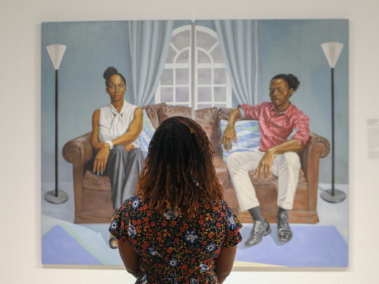 Une personne portant une chemise à fleurs regarde une peinture accrochée au mur de l'exposition Shared Vision au African American Museum in Philadelphia. Le tableau représente deux personnes assises de part et d'autre d'un canapé marron.