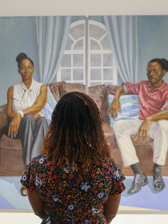 A person wearing a floral shirt looks up at a painting hung on the wall at the Shared Vision exhibit at the African American Museum in Philadelphia. The painting is two people sitting on opposite sides a brown couch.