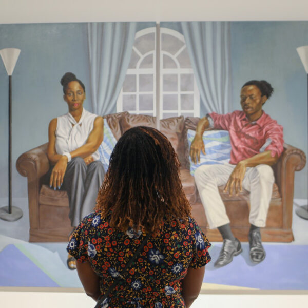A person wearing a floral shirt looks up at a painting hung on the wall at the Shared Vision exhibit at the African American Museum in Philadelphia. The painting is two people sitting on opposite sides a brown couch.