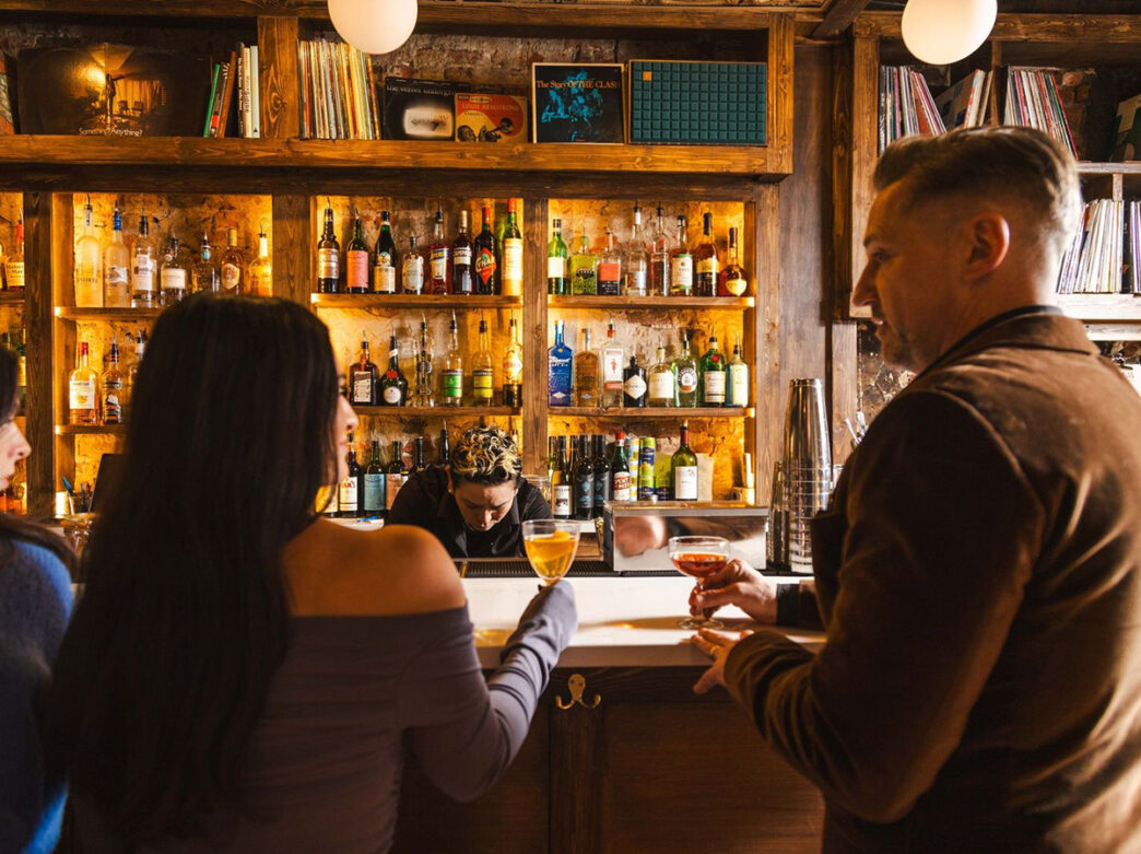 People stand with their backs to the camera while standing at the bar at 48 Record Bar. They hold drinks in cocktail glasses.
