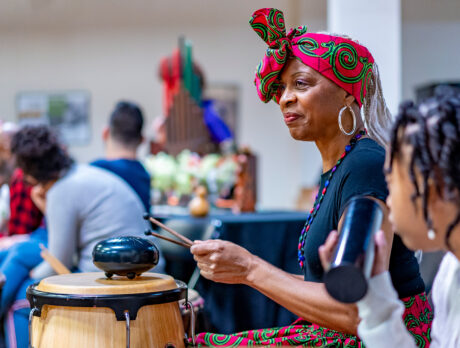 A woman dressed in vibrant red and green traditional attire plays a drum alongside a young child holding a percussion instrument during a Kwanzaa celebration at the African American Museum in Philadelphia.