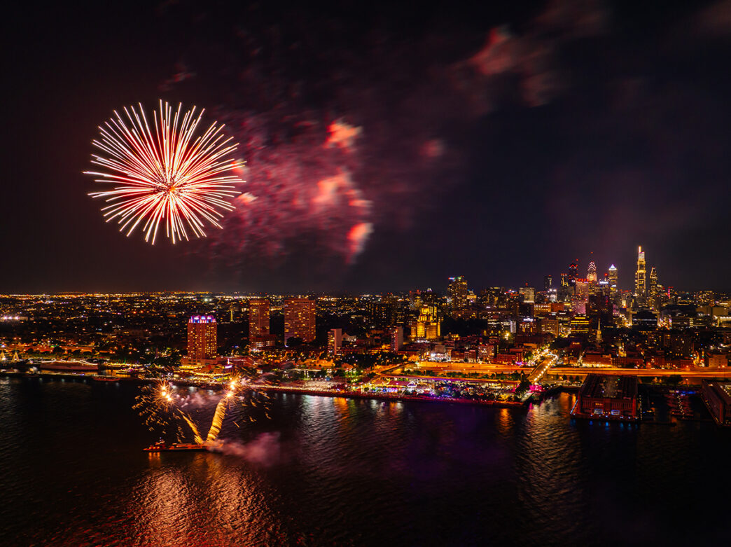 Fireworks explode in the dark sky above the Delaware River. The Philadelphia skyline and Penn's Landing are seen in the image.