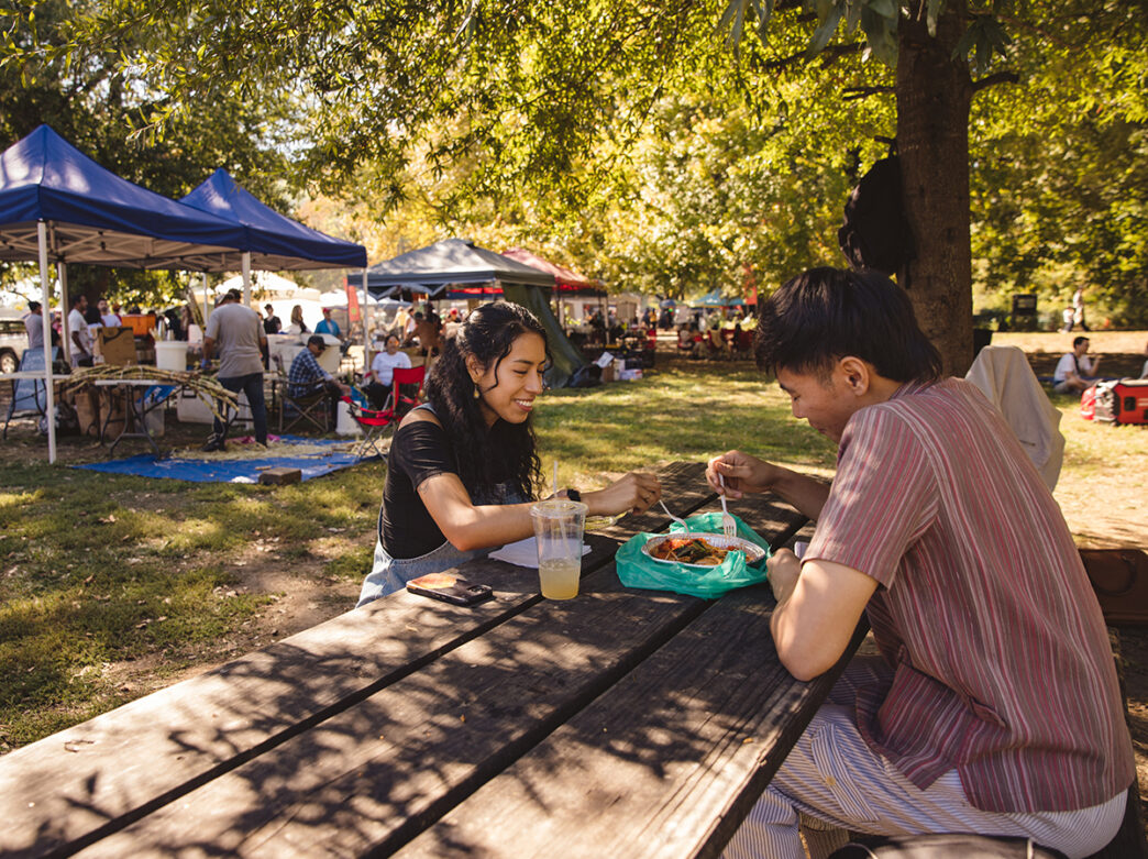 Two people sit across from each other at a picnic table while sharing a meal at the Southeast Asian Market in Philadelphia. Vendor tents can be seen behind the couple.