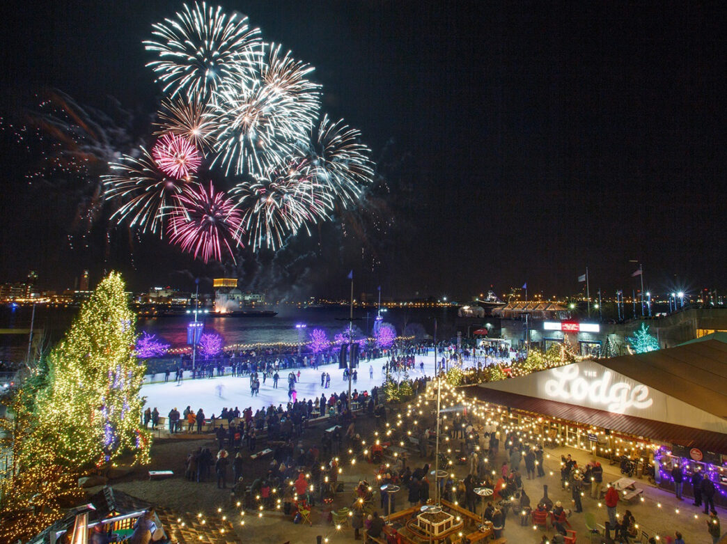 Fireworks explode in the dark sky over the Delaware River and Penn's Landing. People ice skate and watch the fireworks from Blue Cross Riverrink Winterfest.