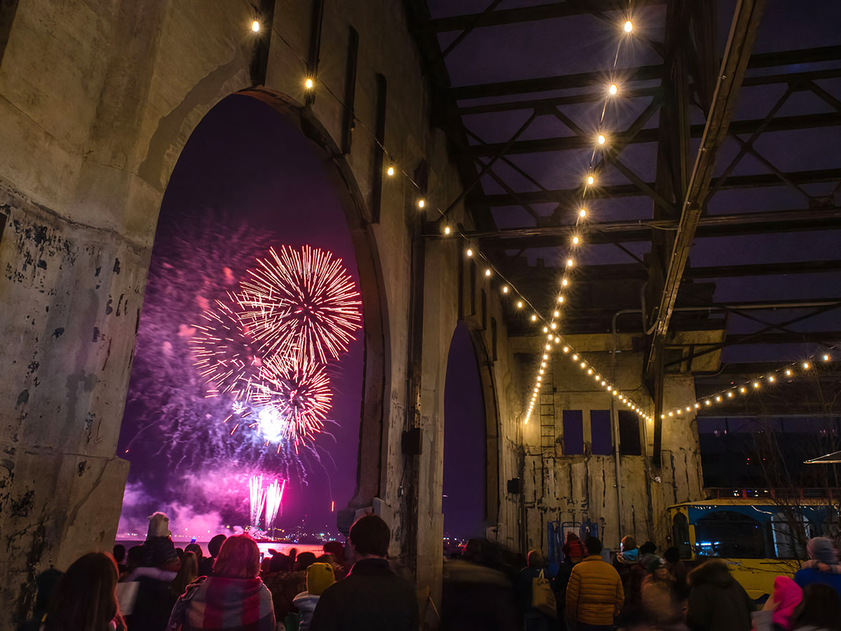 A crowd of people watching the New Year's Eve fireworks from Cherry Street Pier, beneath a covered roof and overlooking the water