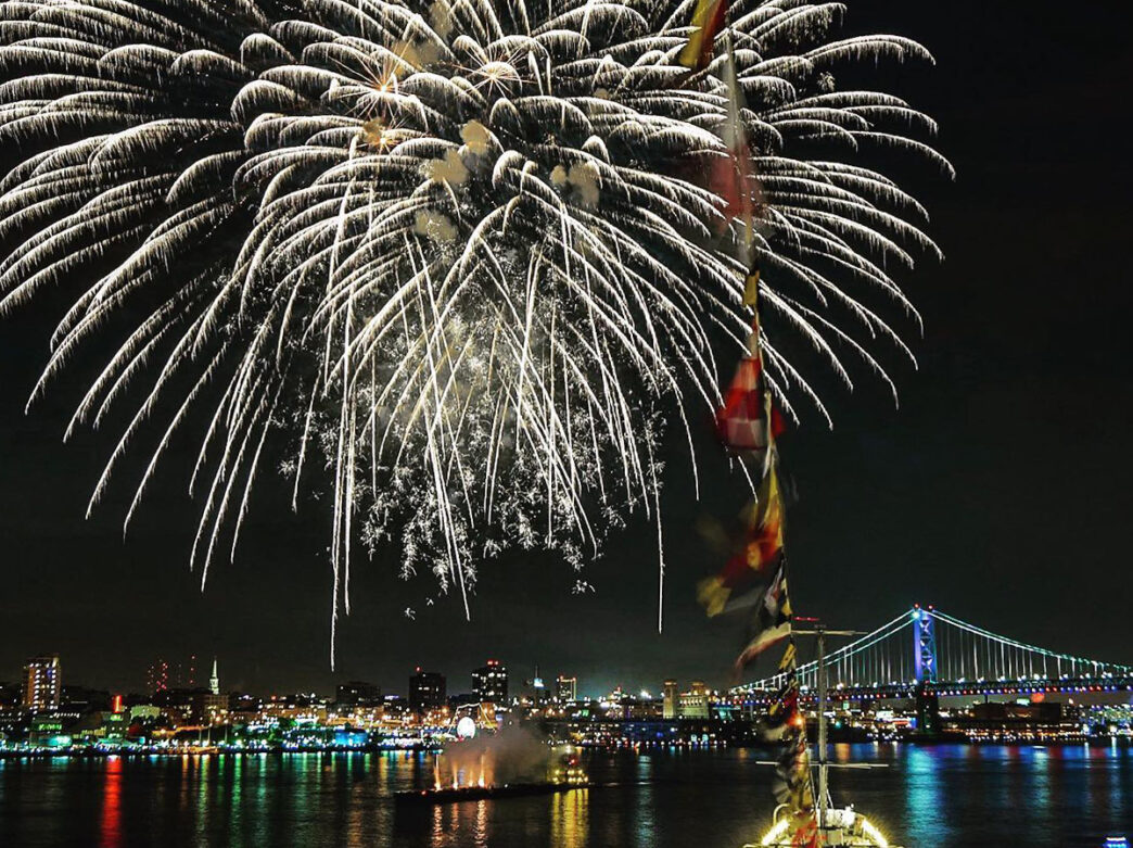 A white firework explodes in the dark sky above the Delaware River in Philadelphia. The Benjamin Franklin Bridge and city skyline are seen in the background.