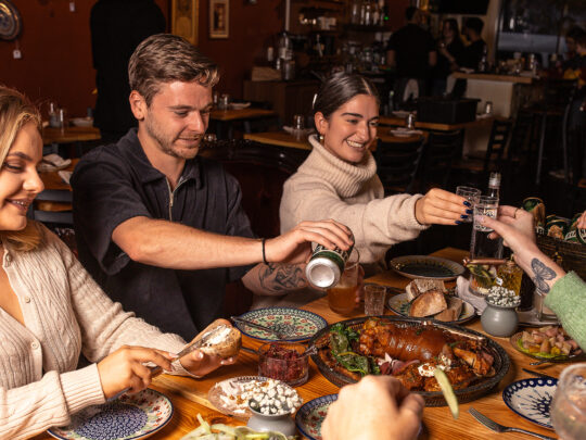 People sit at a dining table, toast glasses, pour beers and spread butter on bread at Little Walter's. Various food items are displayed on the table.