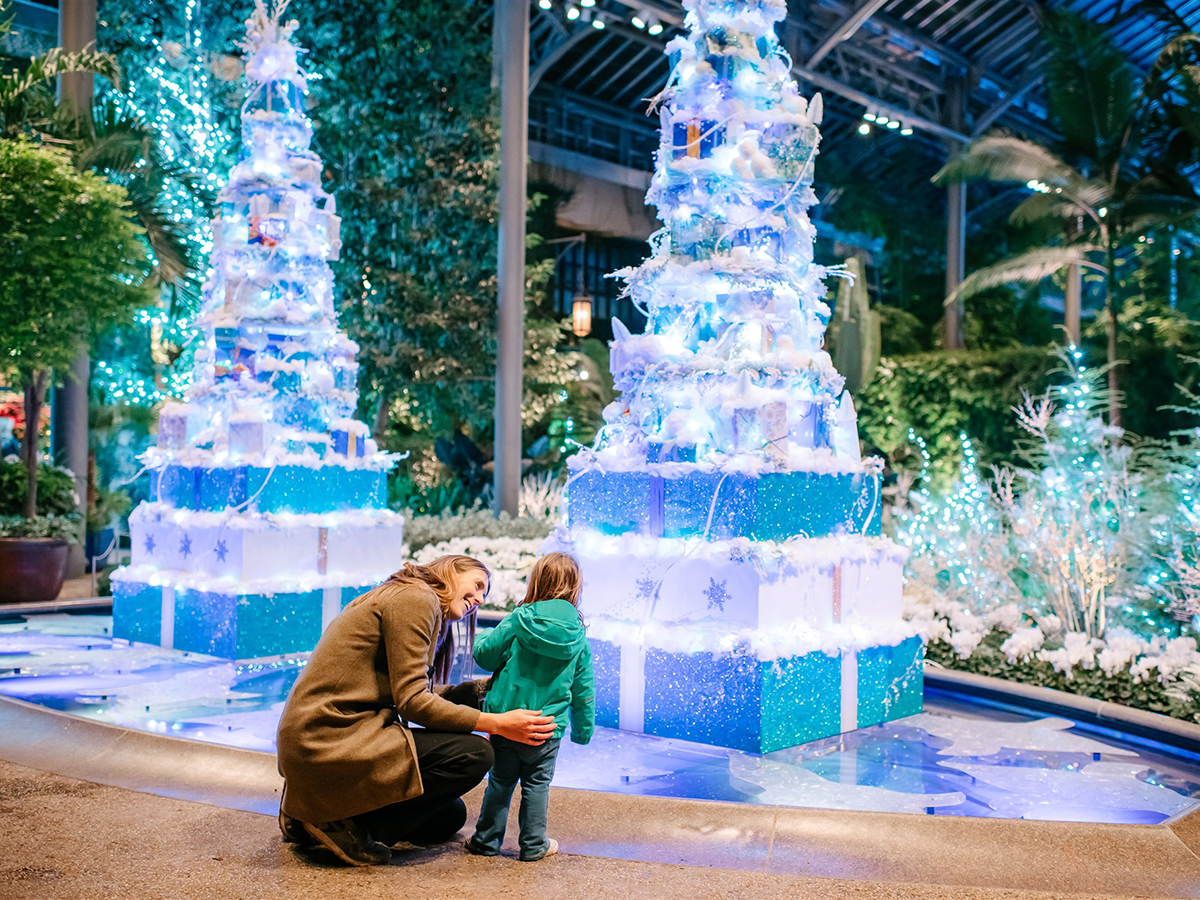 Uma pessoa se agacha ao lado de uma criança e sorri enquanto visita o A Longwood Christmas. O Conservatório Leste está decorado com torres de presentes azuis e brancos brilhantes.