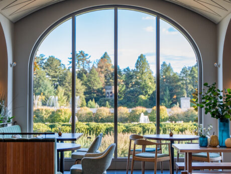 Empty tables and chairs are set up in the 1906 dining room at Longwood Gardens. Floor to ceiling windows overlook gardens and fountains.
