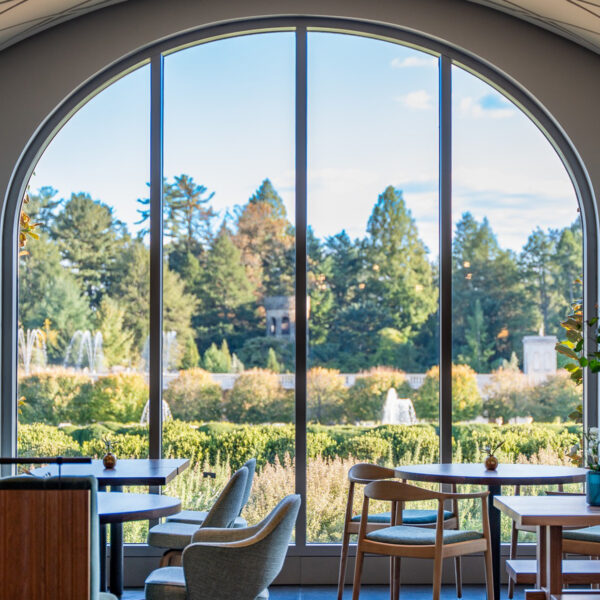 Empty tables and chairs are set up in the 1906 dining room at Longwood Gardens. Floor to ceiling windows overlook gardens and fountains.
