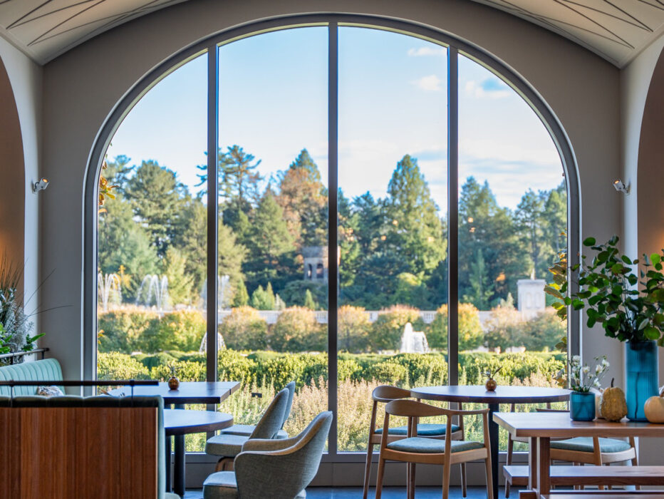 Empty tables and chairs are set up in the 1906 dining room at Longwood Gardens. Floor to ceiling windows overlook gardens and fountains.
