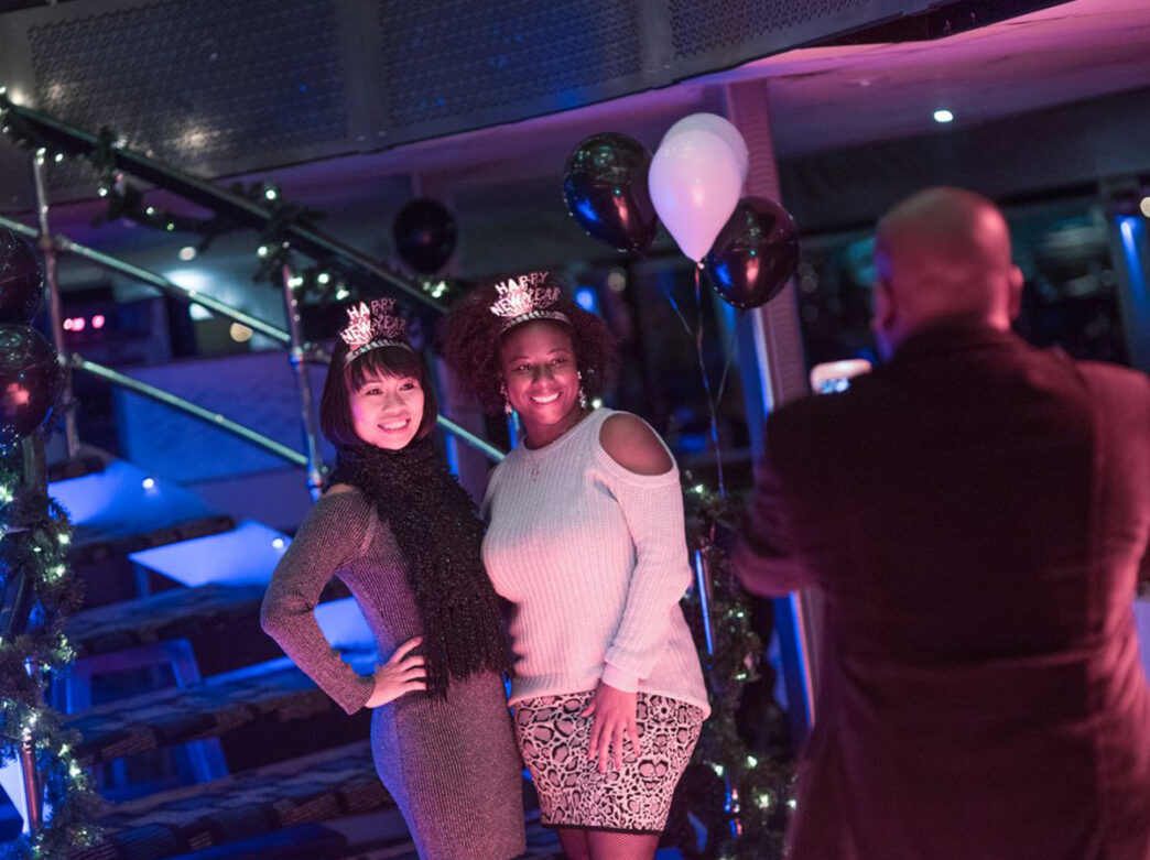 Two people pose for a photo while wearing Happy New Year headbands. Behind them is a staircase decorated with garland and balloons.