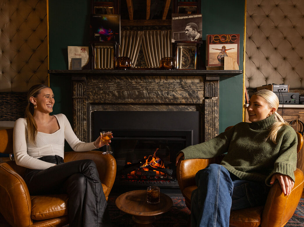 Two women sitting in armchairs by a fireplace, laughing and sipping cocktails. Records are on the wall