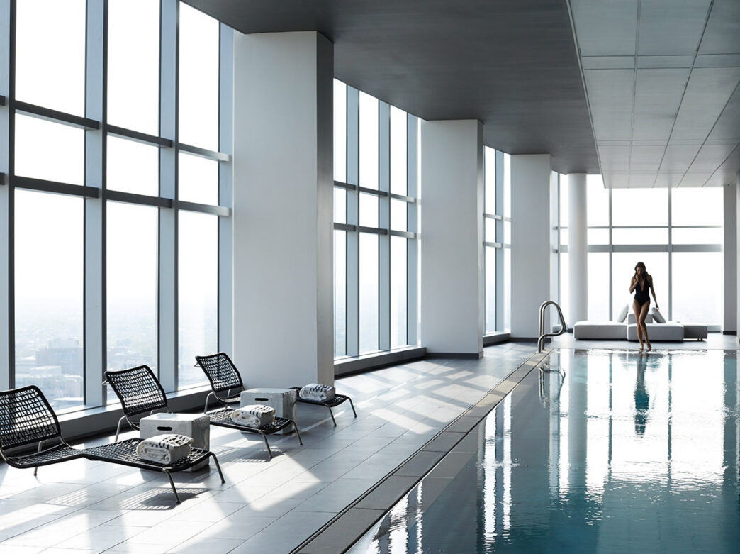 Uma mulher em um maiô preto caminha pela borda da piscina elegante no AKA University City.
