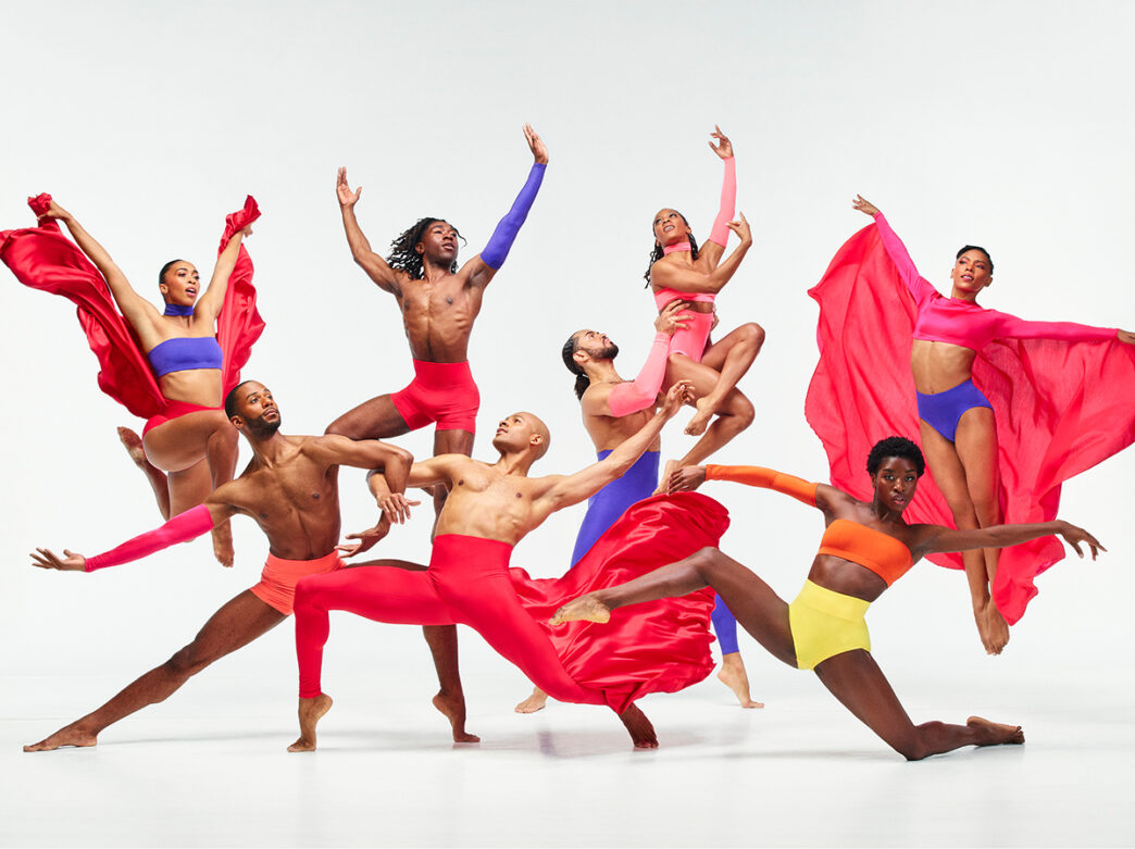 Members of the Alvin Ailey American Dance Theater pose for a group photo. They all wear pink, yellow and blue outfits and pose in various dance poses, many with their arms extended to their sides or above their heads.