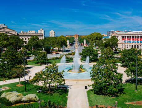 A vibrant view of the Benjamin Franklin Parkway in Philadelphia, featuring the Swann Memorial Fountain in the foreground with its striking water jets surrounded by lush greenery.