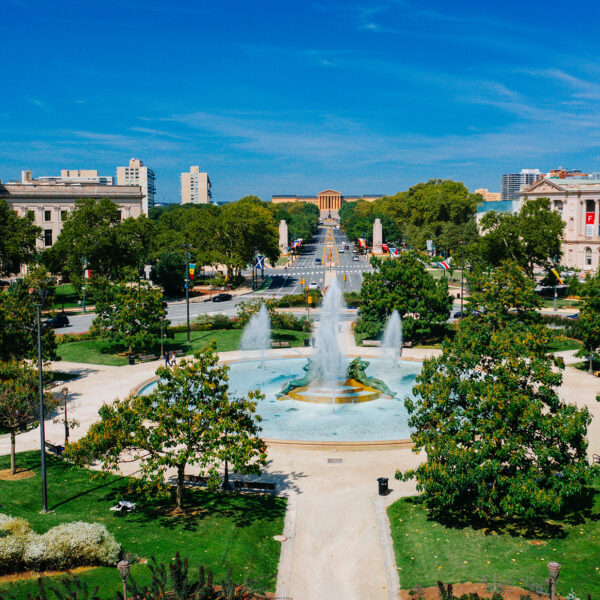 A vibrant view of the Benjamin Franklin Parkway in Philadelphia, featuring the Swann Memorial Fountain in the foreground with its striking water jets surrounded by lush greenery.