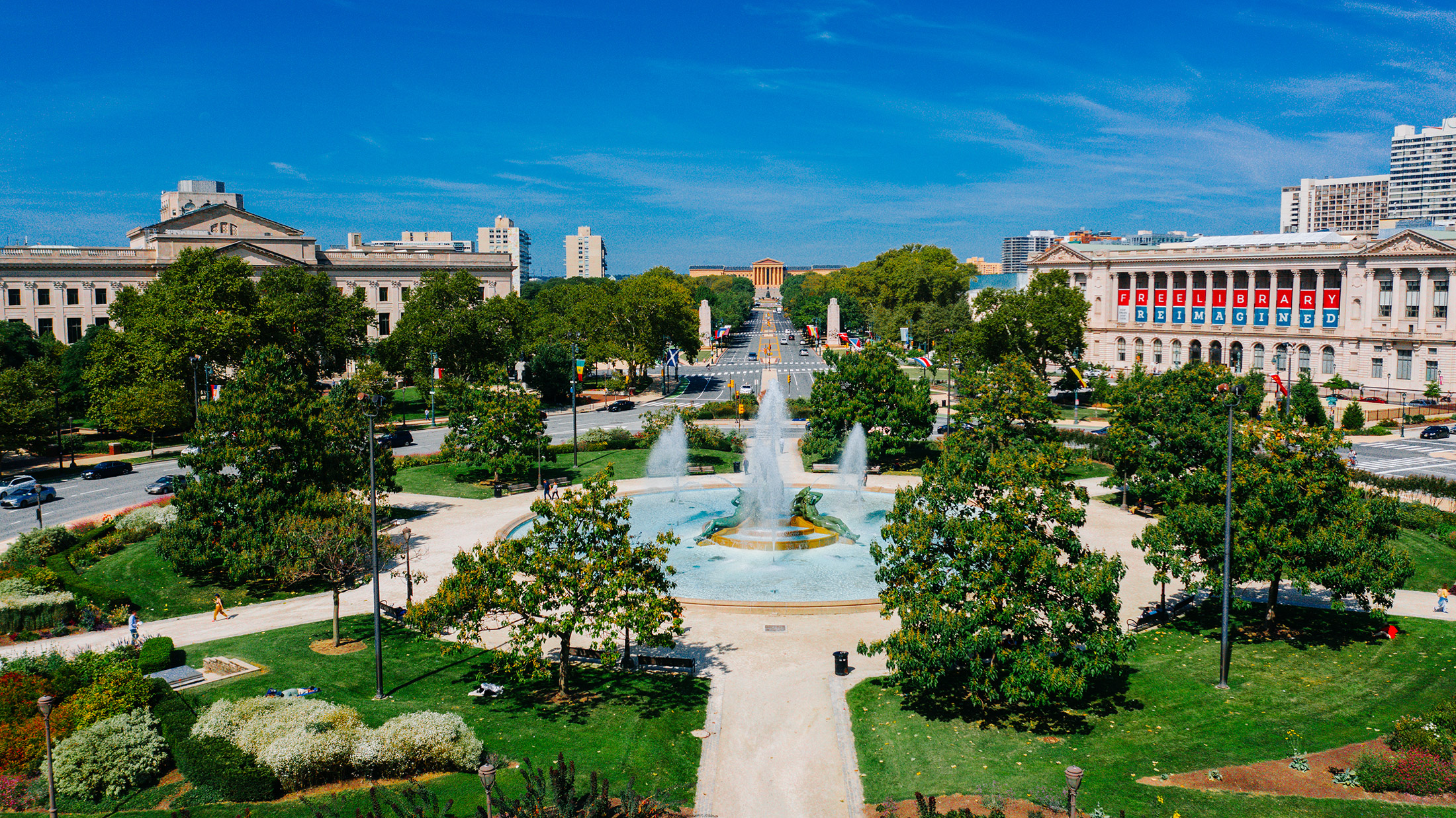 A vibrant view of the Benjamin Franklin Parkway in Philadelphia, featuring the Swann Memorial Fountain in the foreground with its striking water jets surrounded by lush greenery.