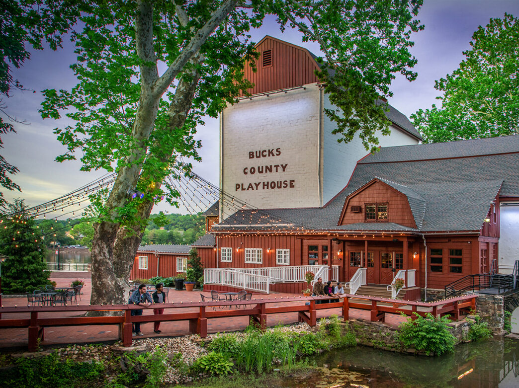 The exterior of a red barn structure that has been converted to the Buck County Playhouse. People stand on a red brick patio in front of the playhouse.