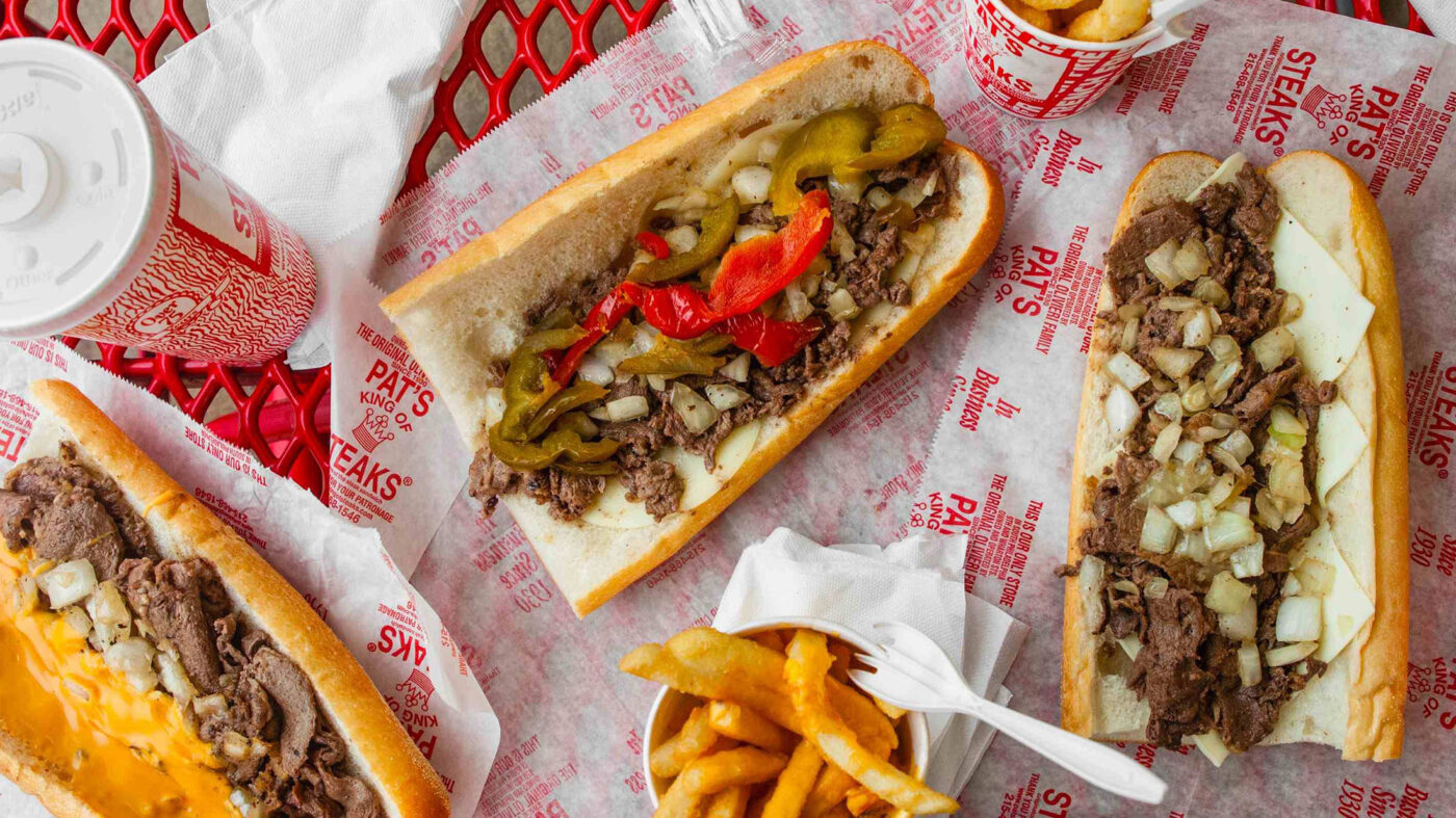 Cheesesteaks, cheese fries and soda cups laid out on a red table.