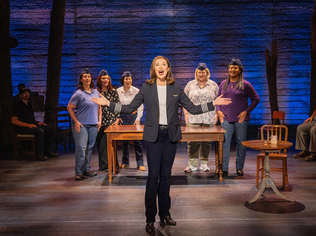An actress dressed as a pilot holds her arms out and sings while on stage during a production of Come From Away. Five actresses stand behind a table in the background.