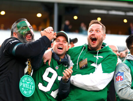 Three Philadelphia Eagles fans hug each other and celebrate while watching a game. One fan's face is painted white and green and the other two wear green Eagles attire.