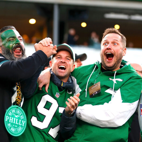 Three Philadelphia Eagles fans hug each other and celebrate while watching a game. One fan's face is painted white and green and the other two wear green Eagles attire.