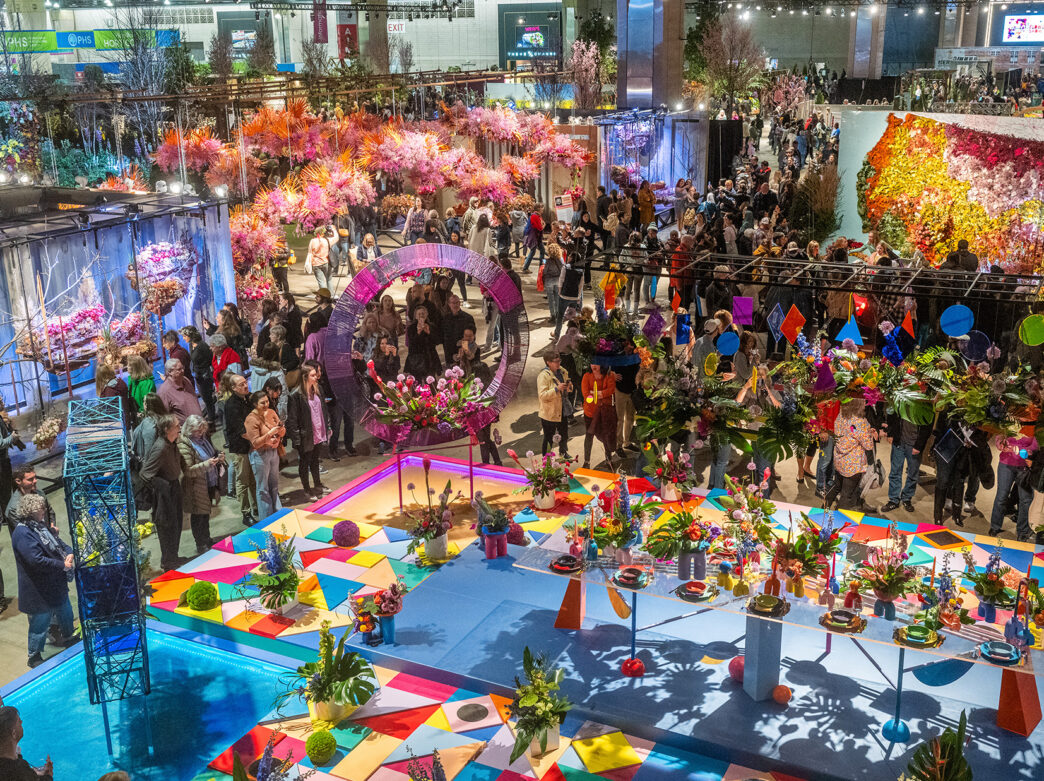 An aerial view of the Philadelphia Flower Show exhibit hall. People walk around and observe the colorful floral displays.