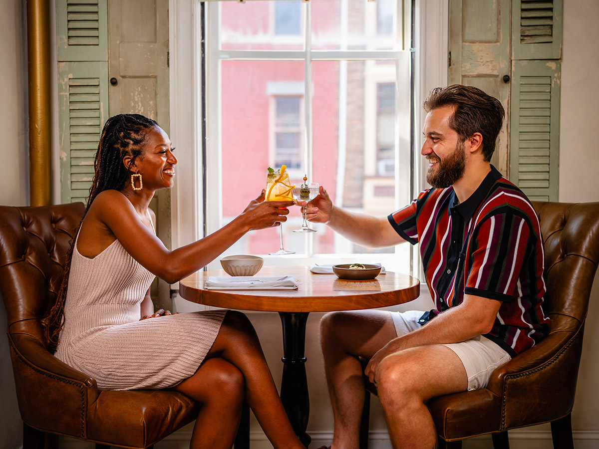 A couple, sitting at a wooden table in front of window in cozy, leather chairs, toast their cocktails.