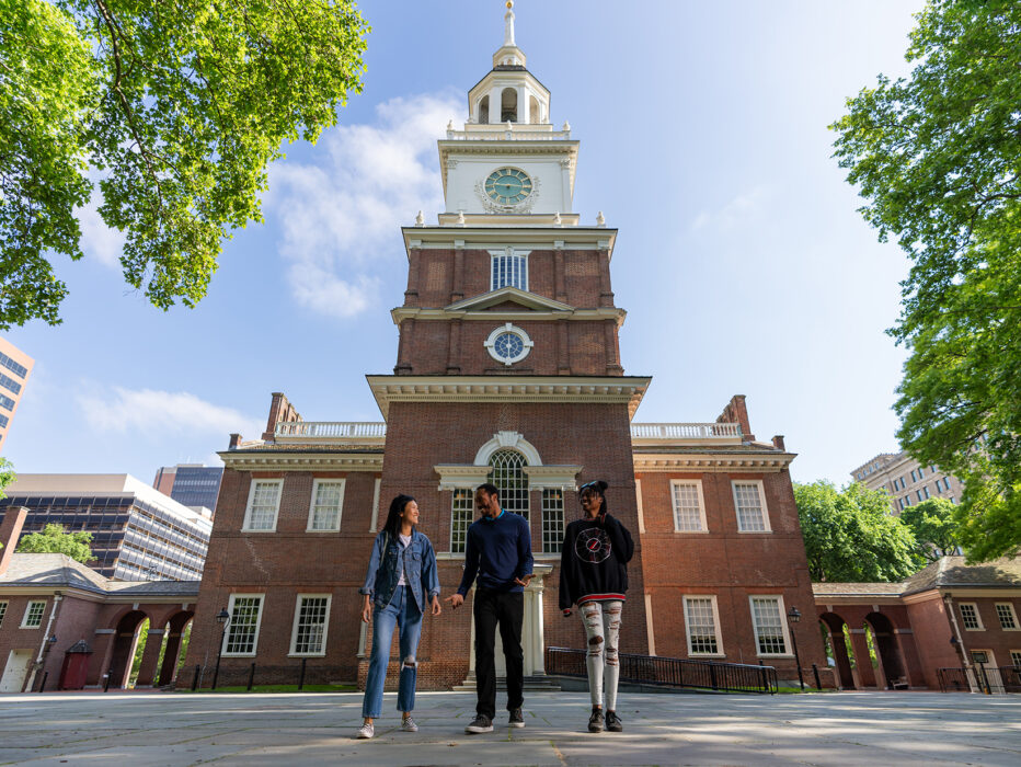 Three people walk with the back of Philadelphia's Independence Hall in the background.