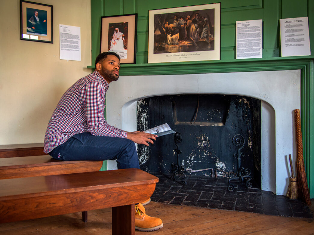 A man sits on a bench in front of a fireplace at the Johnson House in Philadelphia. He holds a pamphlet and looks into the distance. Documents and photos are displayed on the mantle of the fireplace.